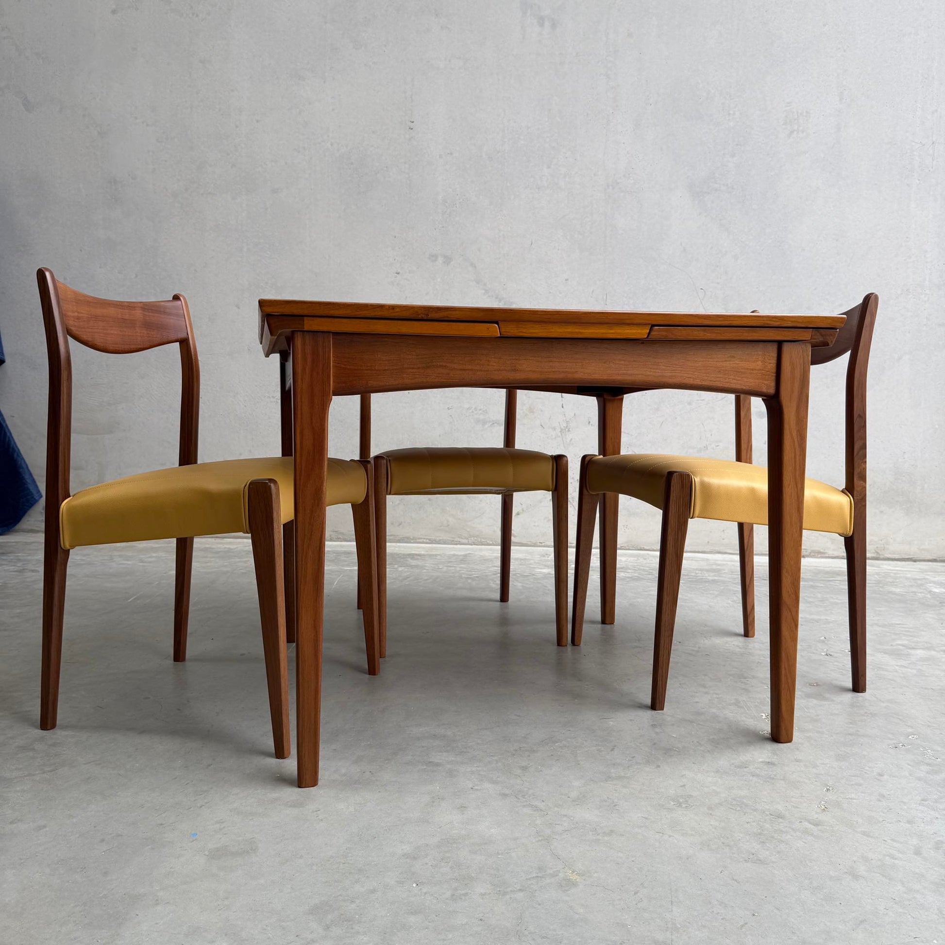 A Fully Restored Fler 64 Square Extension Table (Rediscover) with angled legs is paired with three matching chairs featuring curved backs and yellow seats, set against a plain gray wall and concrete floor.