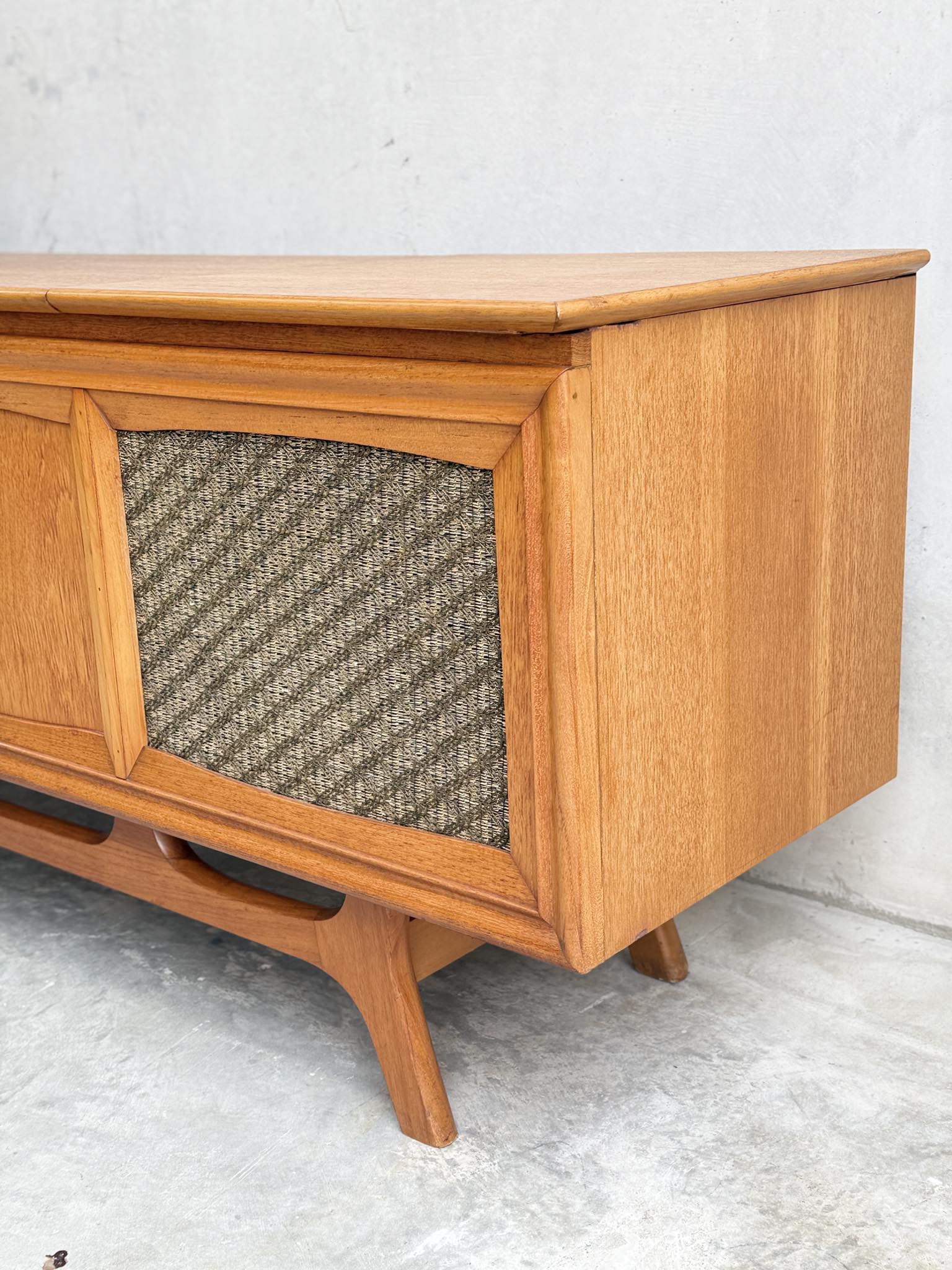 The General Electric Credenza - Spacemaker 250 is a fully converted teak TV/entertainment console with angled legs and a woven textured front, shown against a light concrete wall and floor.