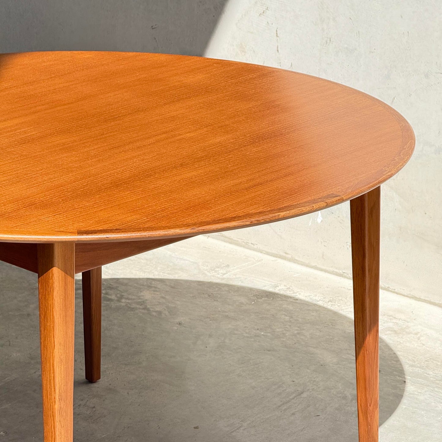 The Fully Restored Noblett Nordic Round MCM Table (Rediscover) features a lacquer finish and tapered legs, set on a light gray concrete floor with an off-white wall backdrop, partly bathed in sunlight.