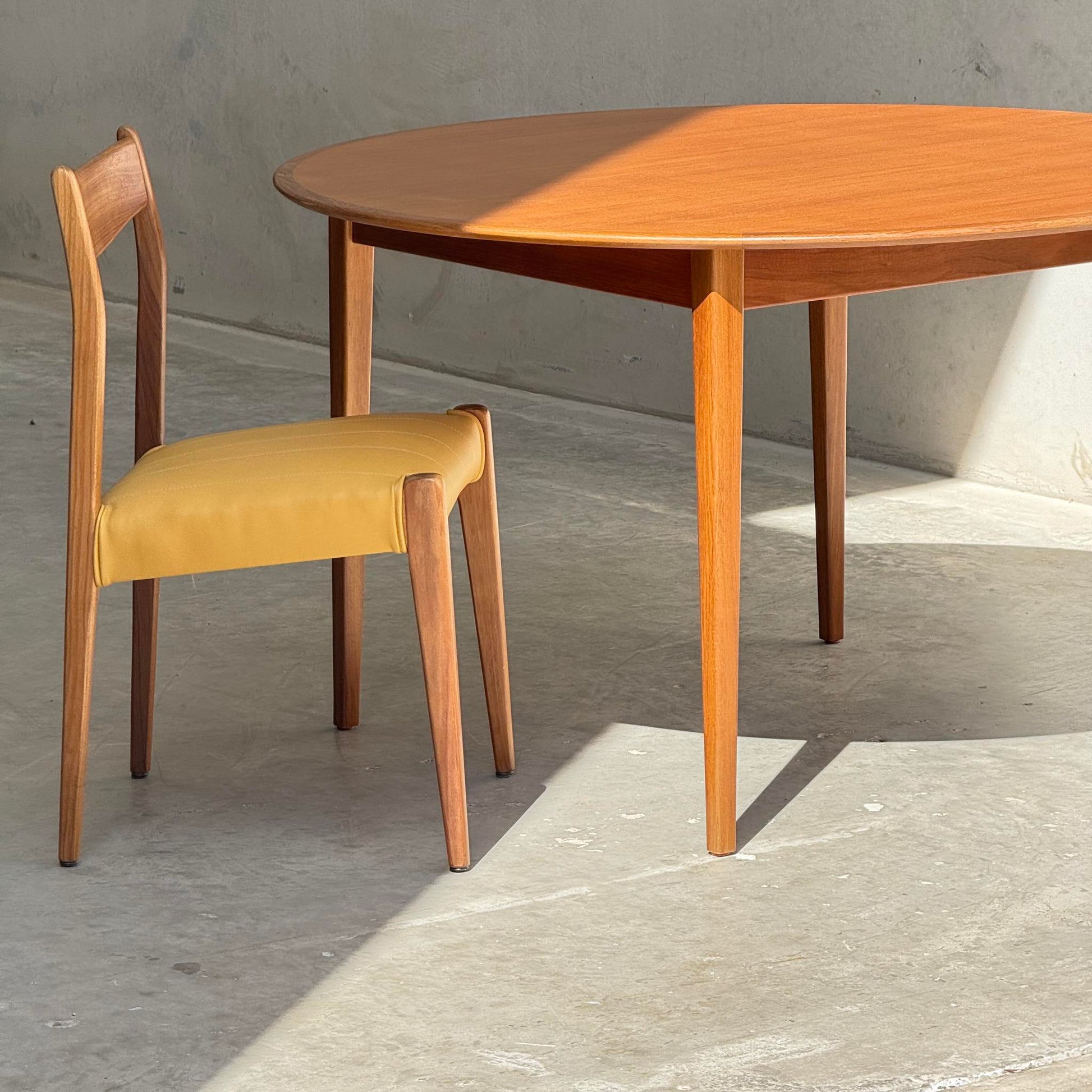 A Fully Restored Noblett Nordic Round MCM Table (Rediscover) stands on a sunlit concrete floor beside a wooden chair with a yellow cushion, casting sharp shadows.