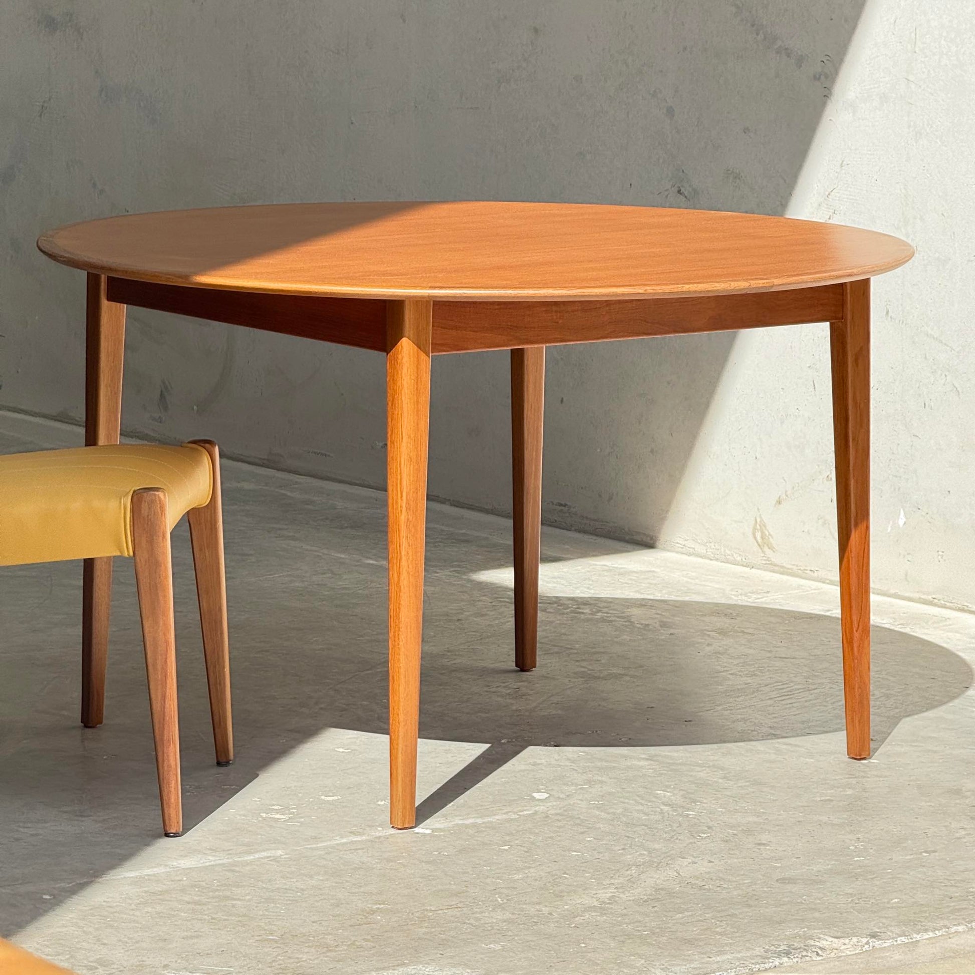 A Fully Restored Noblett Nordic Round MCM Table (Rediscover) with four tapered legs stands in sunlight on a concrete floor, beside a matching wooden chair. Its renewed lacquer finish highlights the timeless mid-century style against the bright wall.