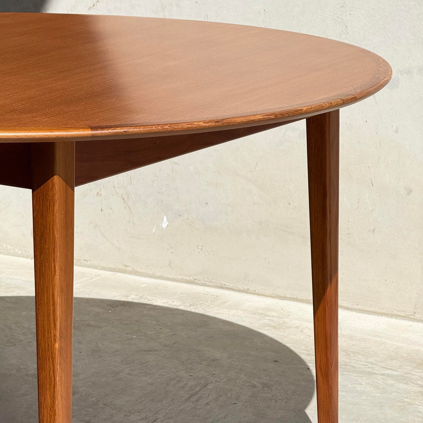 A close-up of the Fully Restored Noblett Nordic Round MCM Table (Rediscover) showcases its sleek lacquer finish and tapered legs, casting a gentle shadow on a sunlit concrete floor beside a light-colored wall.