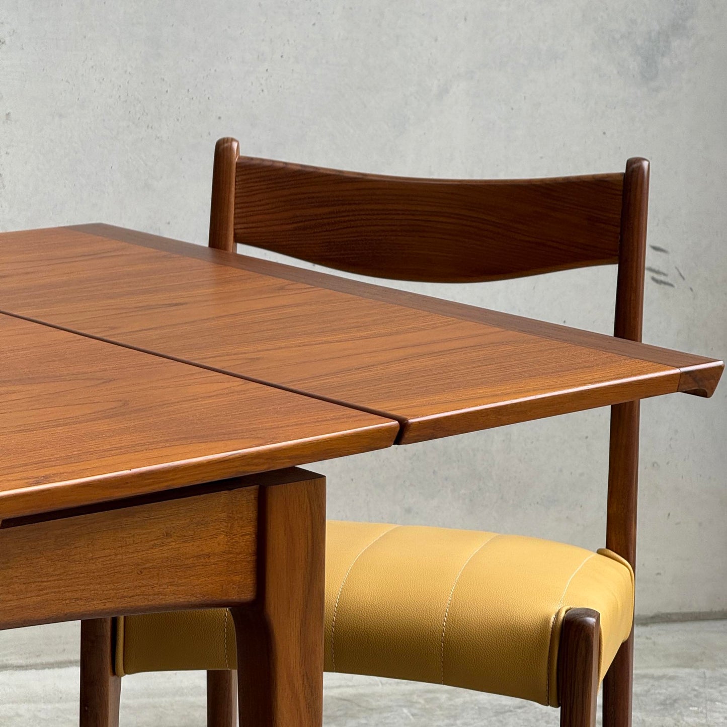 A close-up of the Fully Restored Fler 64 Square Extension Table (Rediscover), paired with a matching wooden chair with a yellow cushioned seat, set before a plain light gray concrete wall.