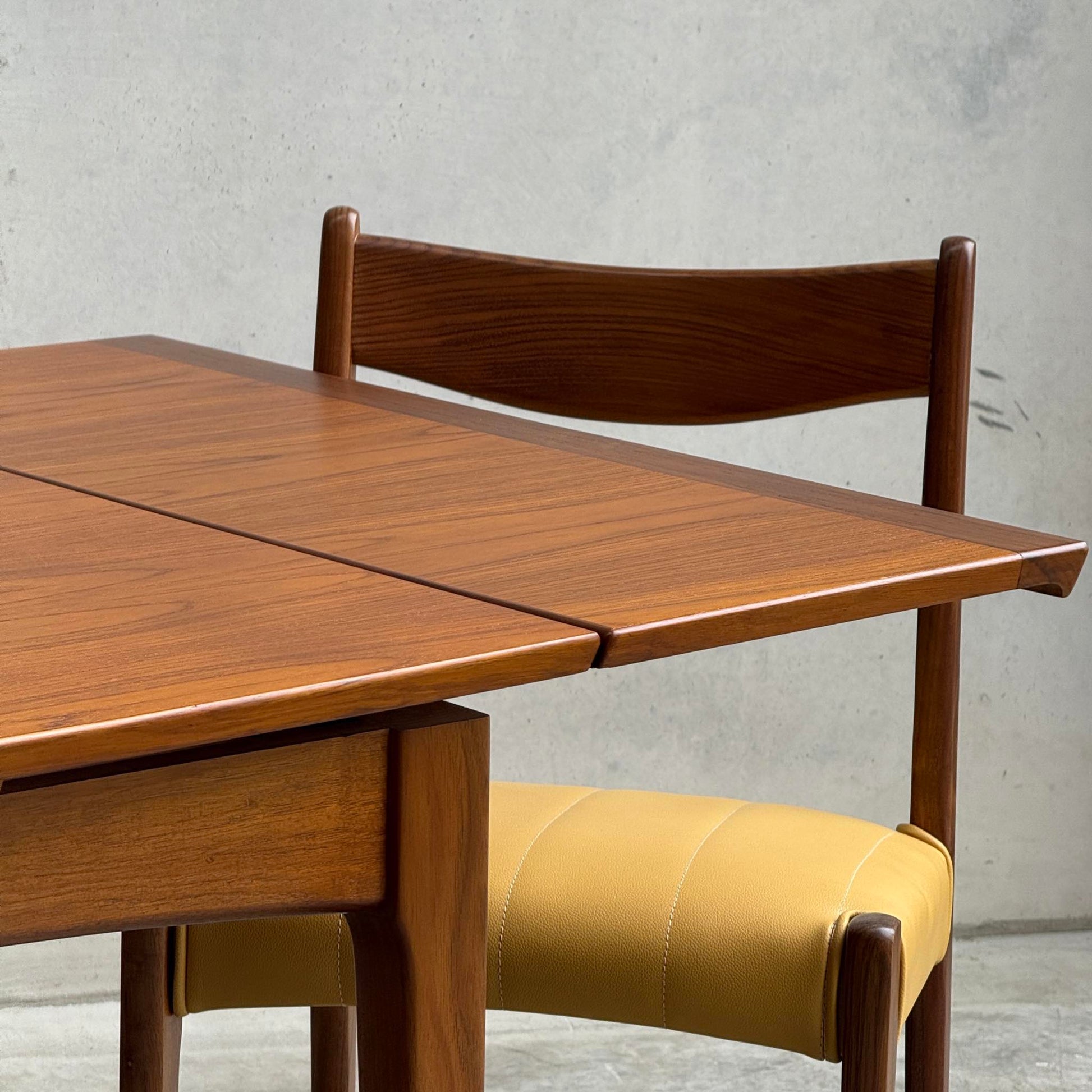 A close-up of the Fully Restored Fler 64 Square Extension Table (Rediscover), paired with a matching wooden chair with a yellow cushioned seat, set before a plain light gray concrete wall.