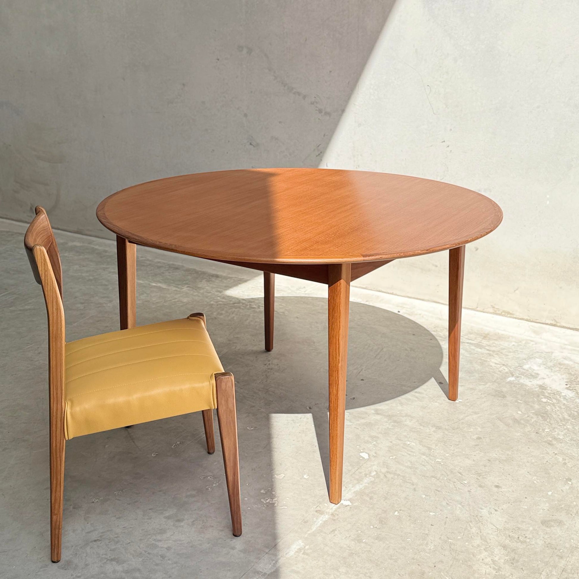 A Fully Restored Noblett Nordic Round MCM Table (Rediscover) with a lacquer finish and tapered legs stands beside a wooden chair with a yellow cushion, sunlit on a light concrete floor against a plain wall.