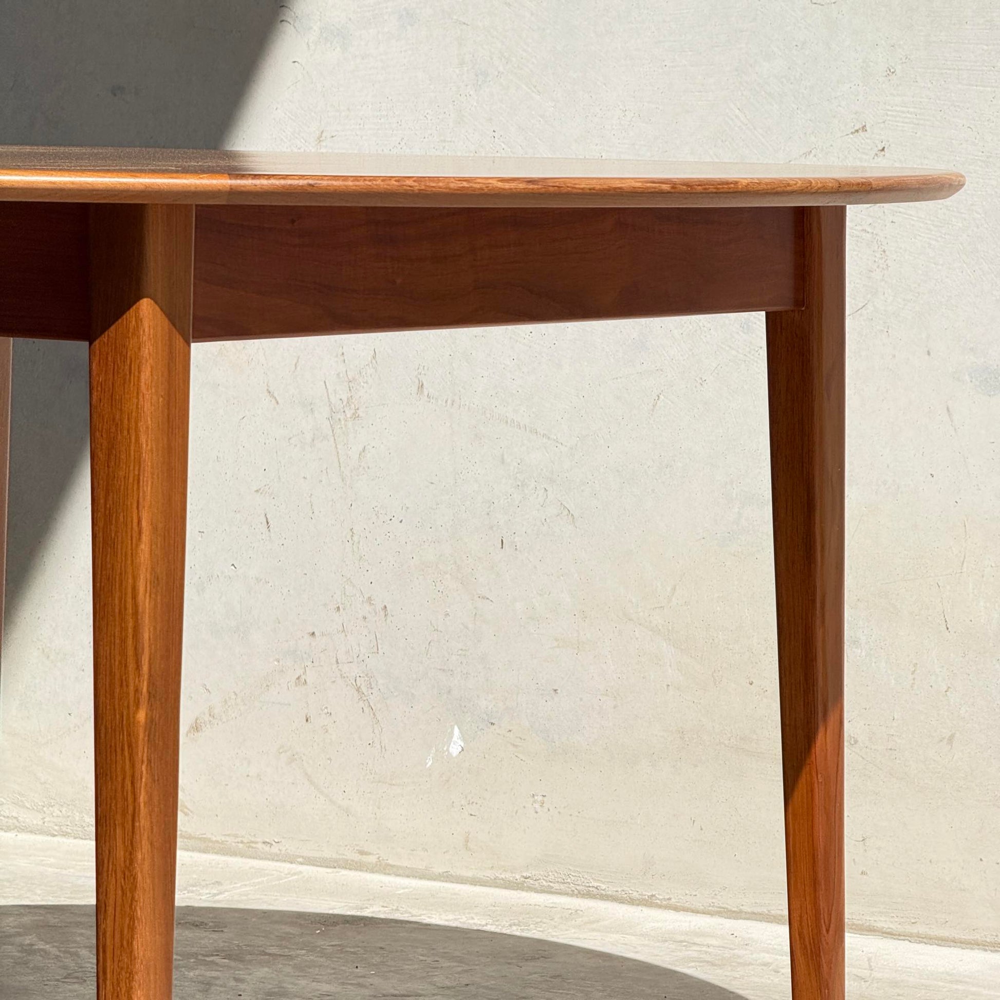 A close-up of the Fully Restored Noblett Nordic Round MCM Table (Rediscover) showcases its tapered legs and smooth edge, with a gleaming lacquer finish set against a textured, light-colored wall in bright sunlight.