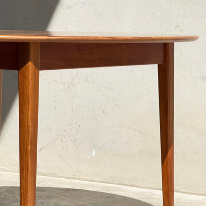 A close-up of the Fully Restored Noblett Nordic Round MCM Table (Rediscover) showcases its tapered legs and smooth edge, with a gleaming lacquer finish set against a textured, light-colored wall in bright sunlight.