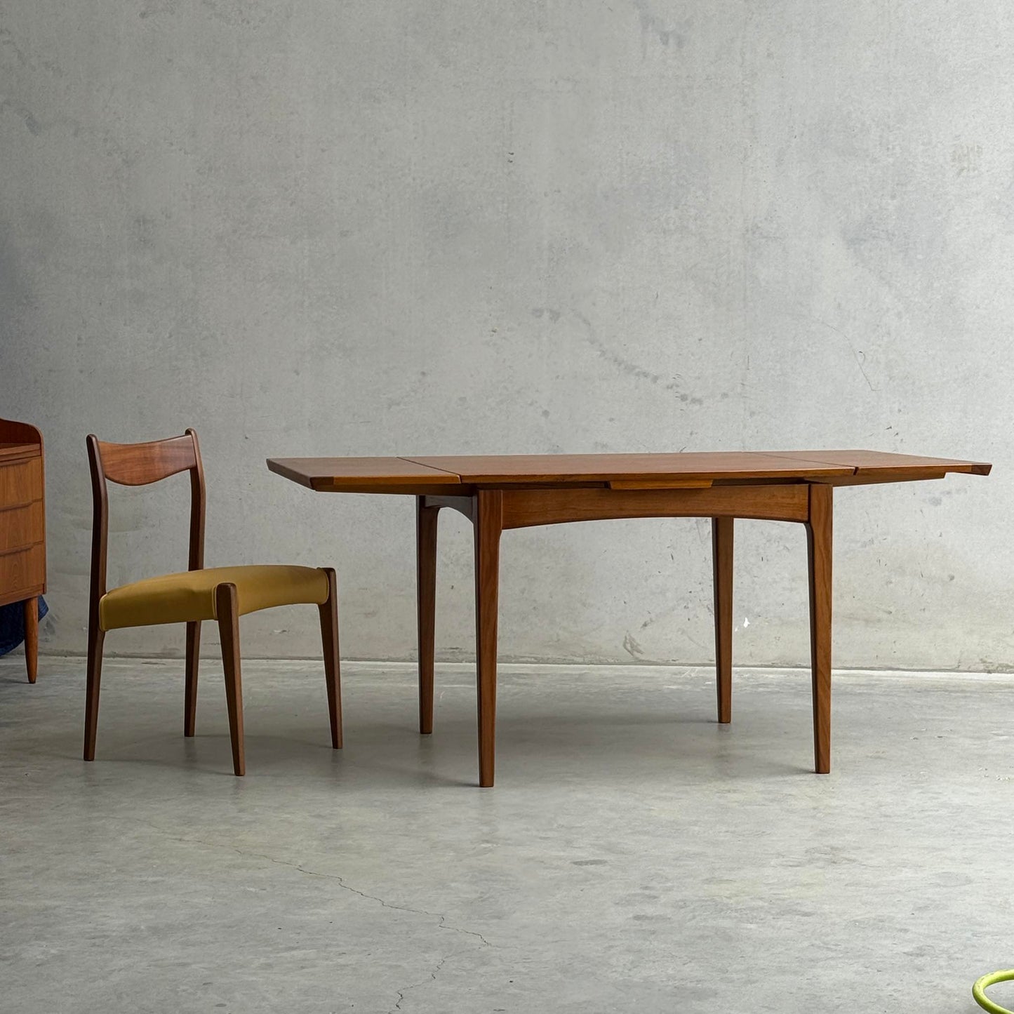 A Fully Restored Fler 64 Square extension Table (Rediscover) with a matching wooden chair featuring a yellow upholstered seat stands out against a gray concrete wall and floor, exemplifying timeless mid-century modern style.