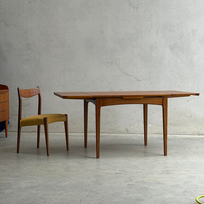 A Fully Restored Fler 64 Square extension Table (Rediscover) with a matching wooden chair featuring a yellow upholstered seat stands out against a gray concrete wall and floor, exemplifying timeless mid-century modern style.