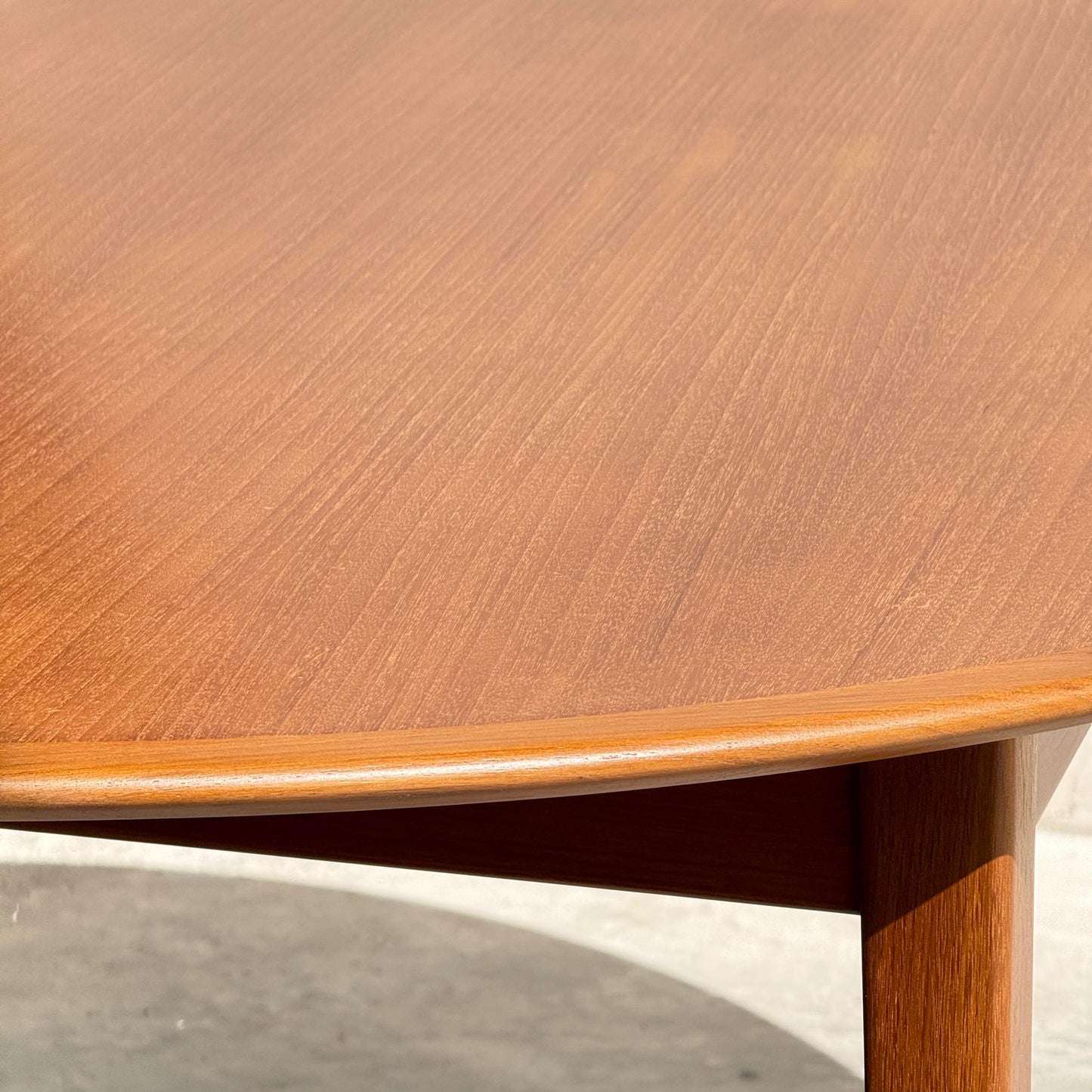 Close-up of the Fully Restored Noblett Nordic Round MCM Table (Rediscover), featuring its renewed lacquer finish and rich wood grain, with sunlight highlighting the detail. A table leg and shadow appear in the background, reflecting its mid-century modern appeal.
