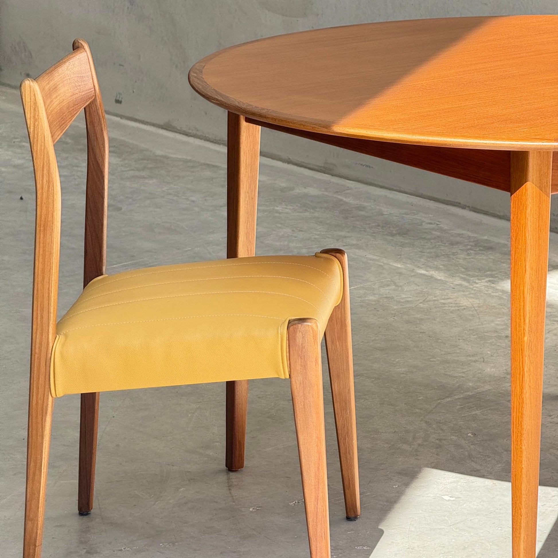 A wooden chair with a yellow cushioned seat sits beside a Fully Restored Noblett Nordic Round MCM Table (Rediscover) on a concrete floor, with sunlight casting soft shadows across the table and floor.