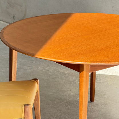 A Fully Restored Noblett Nordic Round MCM Table (Rediscover) with a lacquer finish sits on a gray concrete floor beside a tan cushioned chair, sunlight casting diagonal shadows across its mid-century modern surface.