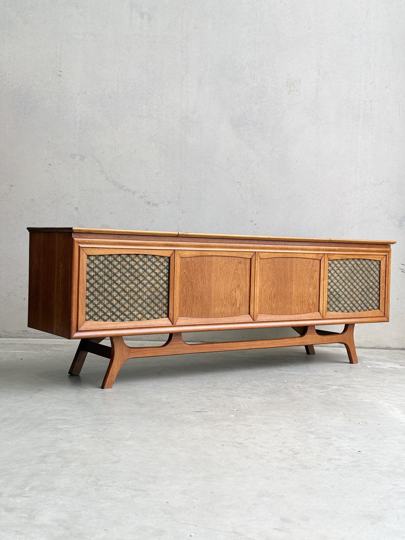 The General Electric Credenza - Spacemaker 250 is a fully converted teak TV and entertainment console with sculpted legs, paneled doors, and patterned speaker grilles, shown against a plain concrete wall and floor.
