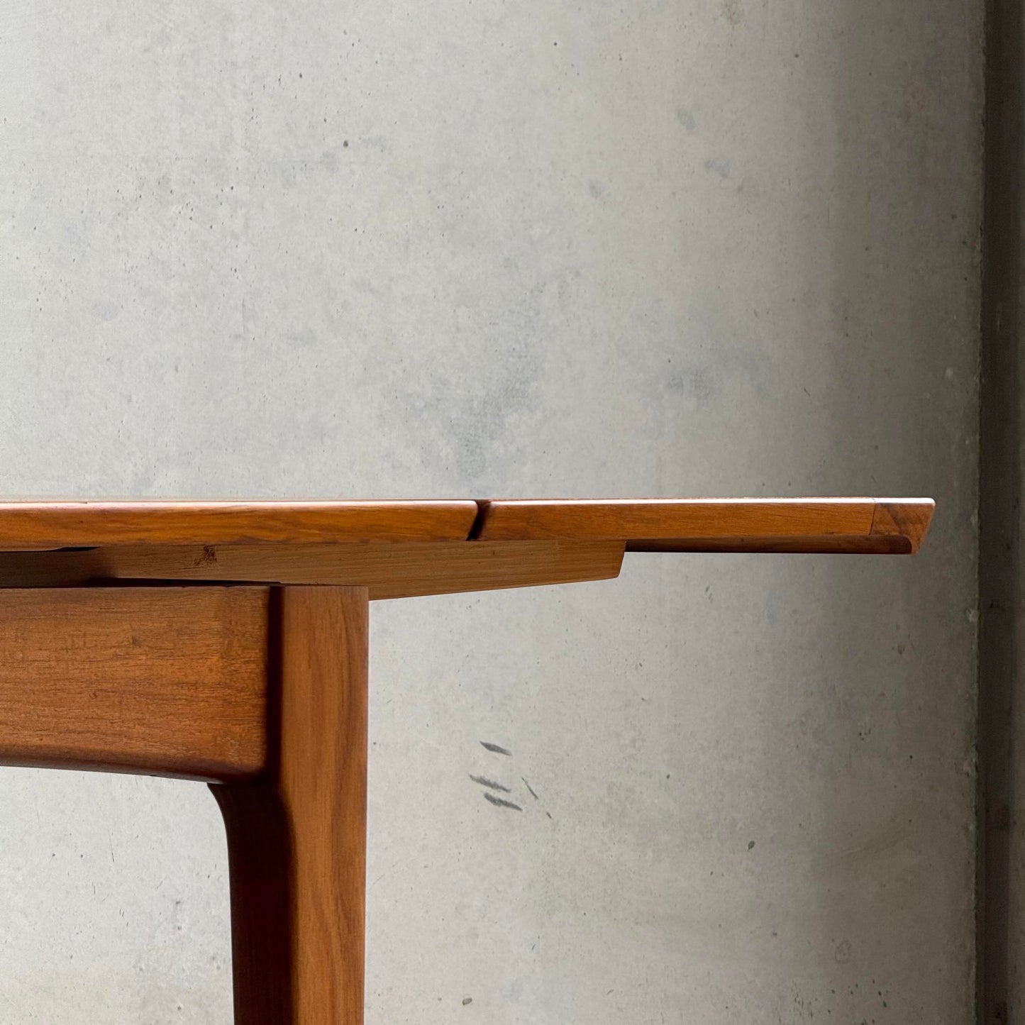 A close-up side view of the Fully Restored Fler 64 Square Extension Table (Rediscover), highlighting its mid-century modern design, clean lines, and warm wood finish against a light, subtly textured background.