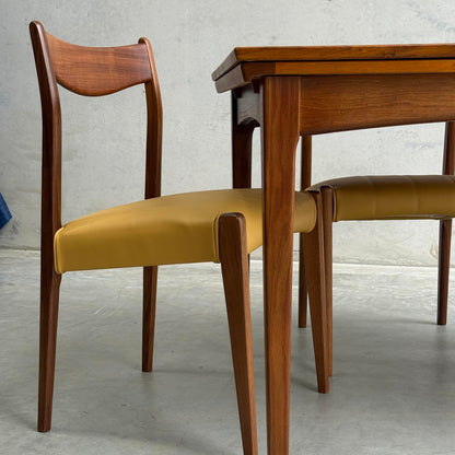 A wooden dining chair with a curved backrest and mustard yellow cushioned seat sits beside the Fully Restored Fler 64 Square Extension Table (Rediscover) on a light gray concrete floor against a plain wall.