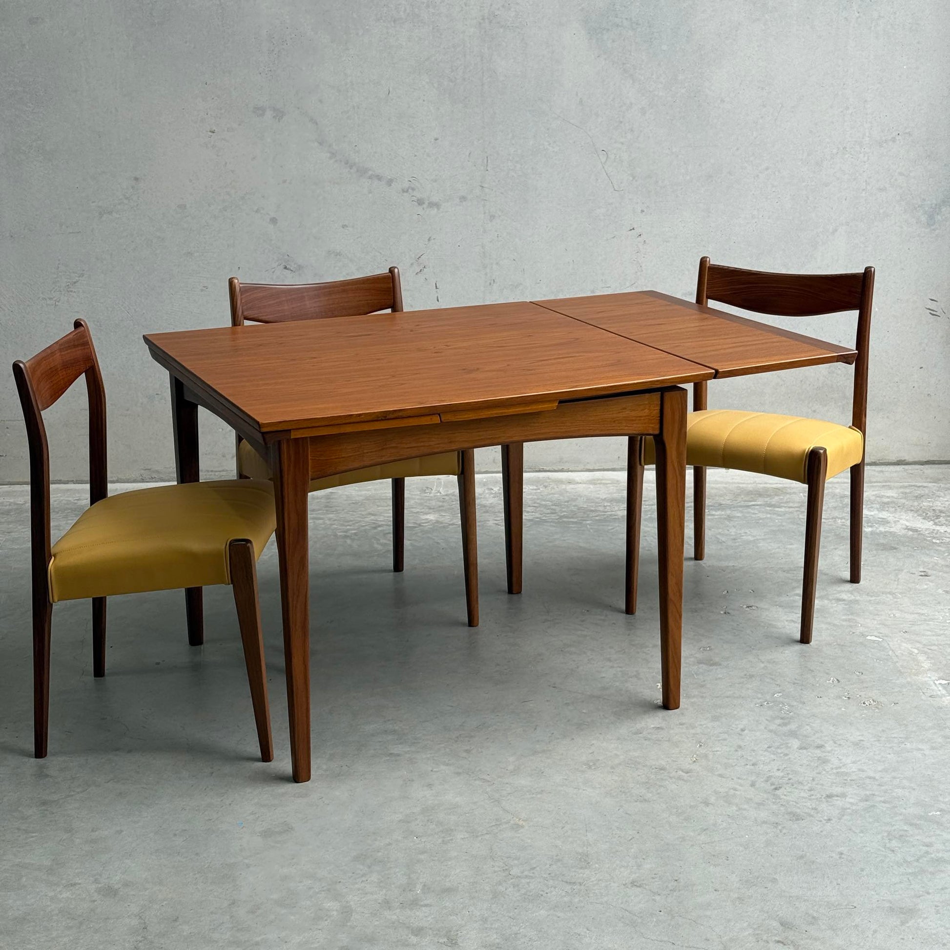A Fully Restored Fler 64 Square Extension Table (Rediscover) with clean lines and three matching chairs, two featuring yellow upholstered seats, is set against a plain gray concrete wall and floor.
