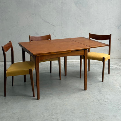 A Fully Restored Fler 64 Square Extension Table (Rediscover) with clean lines and three matching chairs, two featuring yellow upholstered seats, is set against a plain gray concrete wall and floor.