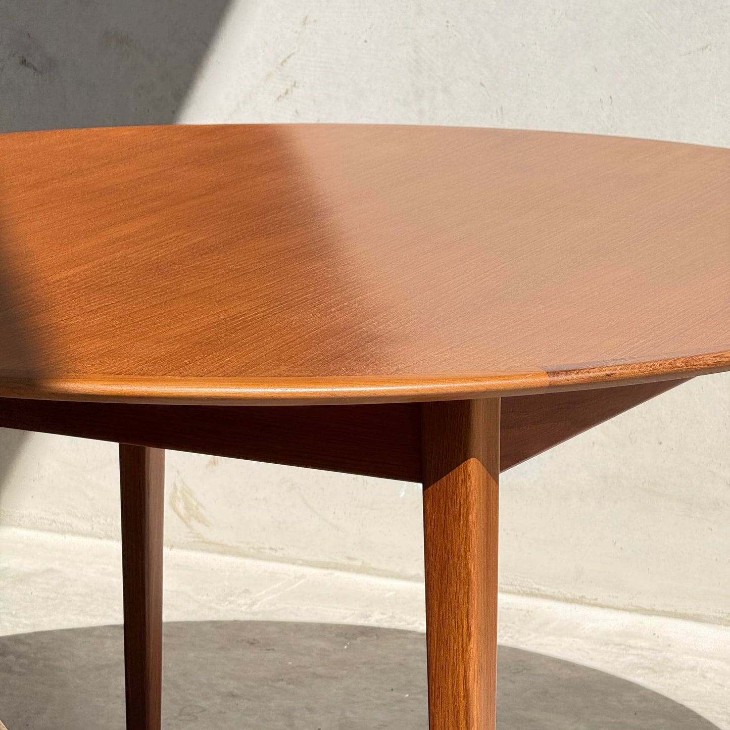 A fully restored Noblett Nordic Round MCM Table (Rediscover) with tapered legs stands in sunlight, its lacquer finish glowing as a diagonal shadow falls across the tabletop against a light wall and concrete floor.