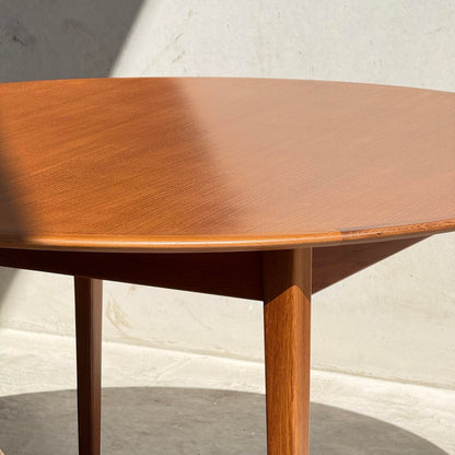 A fully restored Noblett Nordic Round MCM Table (Rediscover) with tapered legs stands in sunlight, its lacquer finish glowing as a diagonal shadow falls across the tabletop against a light wall and concrete floor.