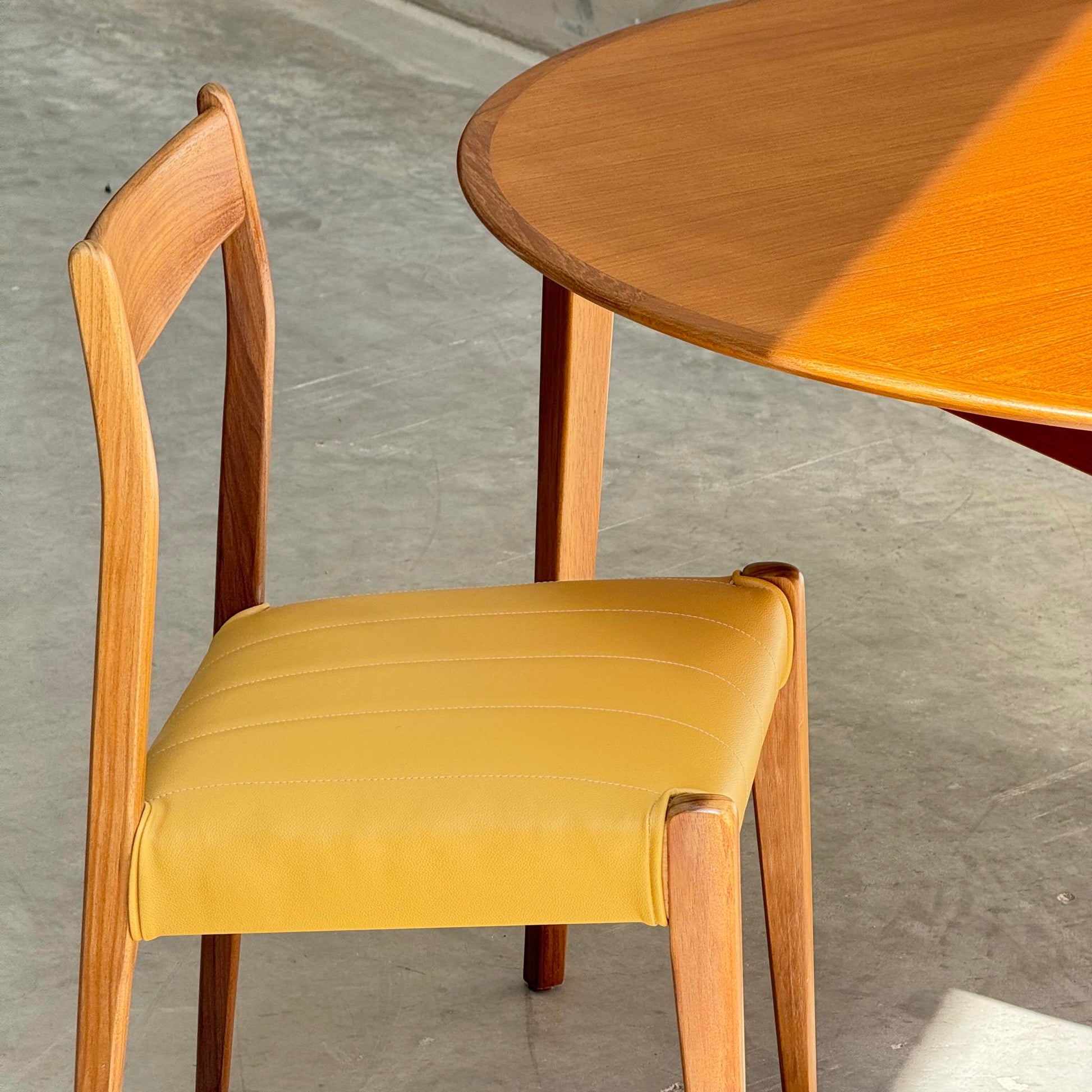A wooden chair with a yellow cushioned seat is beside a Fully Restored Noblett Nordic Round MCM Table (Rediscover) on a concrete floor, illuminated by sunlight.