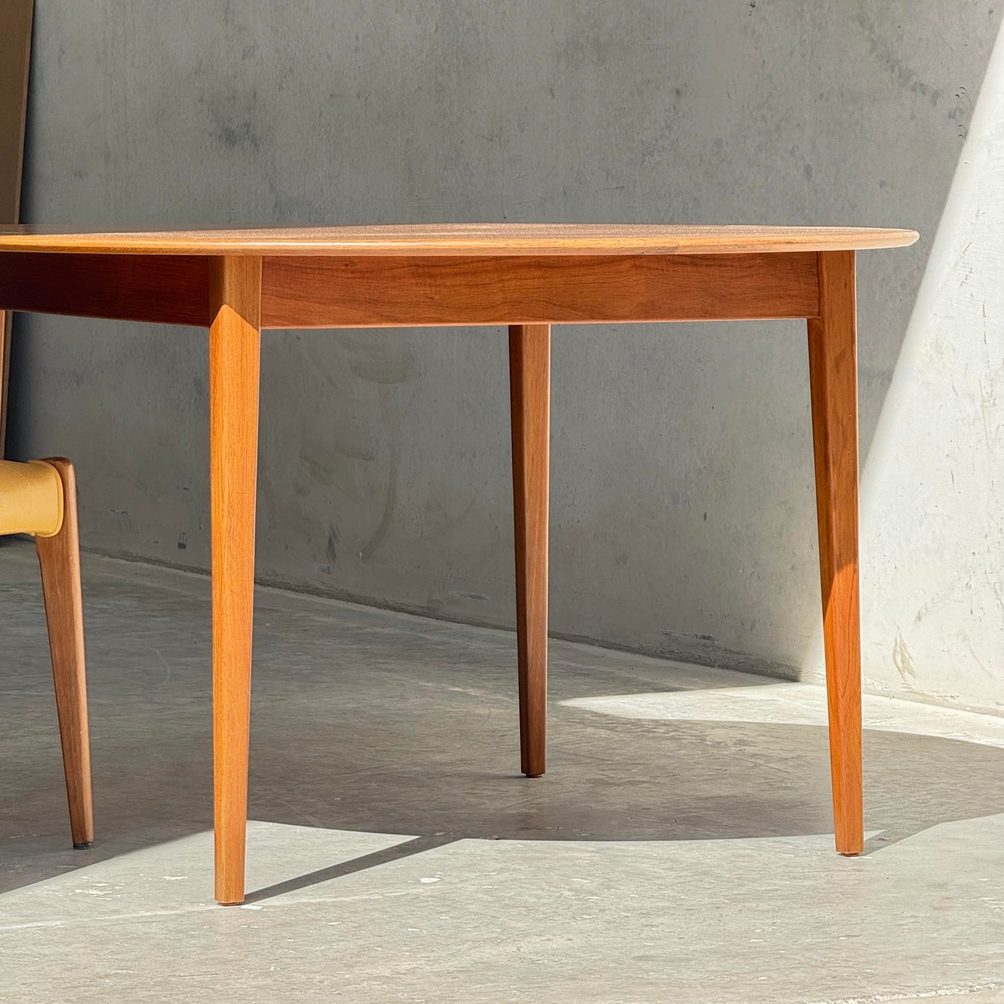 A Fully Restored Noblett Nordic Round MCM Table (Rediscover) with a lacquer finish stands on a concrete floor in a sunlit room; a matching wooden chair is partially visible, completing this mid-century modern dining setting.