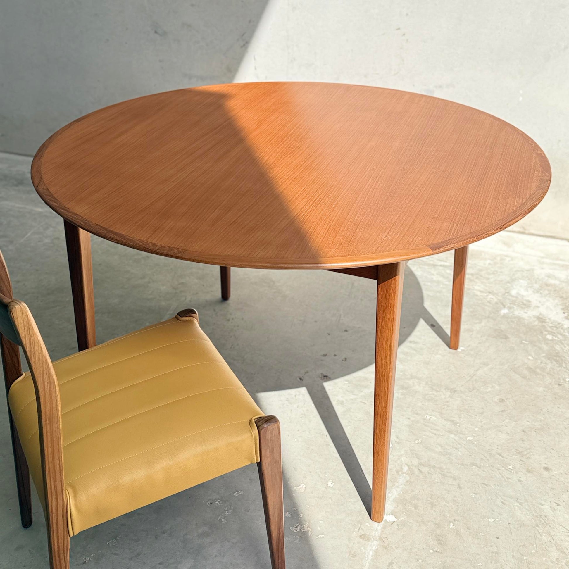 A Fully Restored Noblett Nordic Round MCM Table (Rediscover) with tapered legs stands beside a yellow-cushioned wooden chair, sunlight casting diagonal shadows across its lacquer finish and highlighting its mid-century modern charm.