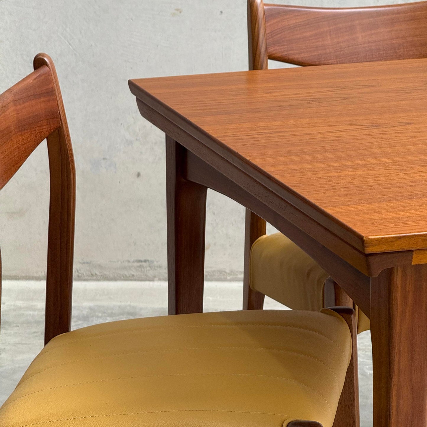 A close-up of the Fully Restored Fler 64 Square Extension Table (Rediscover) with matching wooden chairs featuring tan upholstered seats, set against a plain, light-colored wall and floor.