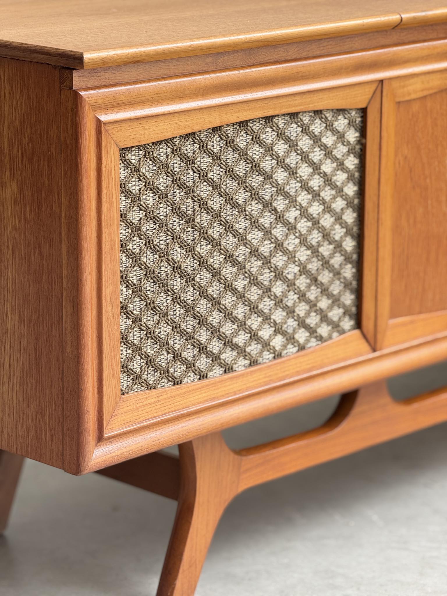 Close-up of a General Electric Credenza Spacemaker 250, a fully converted teak TV/entertainment console with mid-century modern design, featuring book-matched teak, angled legs, and a textured fabric panel on a light floor.