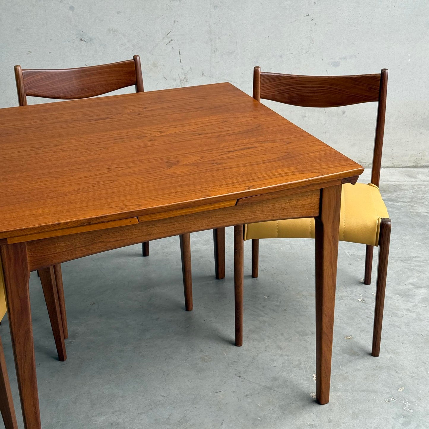 A Fully Restored Fler 64 Square Extension Table (Rediscover) with tapered legs is paired with three matching wooden chairs featuring yellow cushions, set against a plain gray wall and concrete floor.
