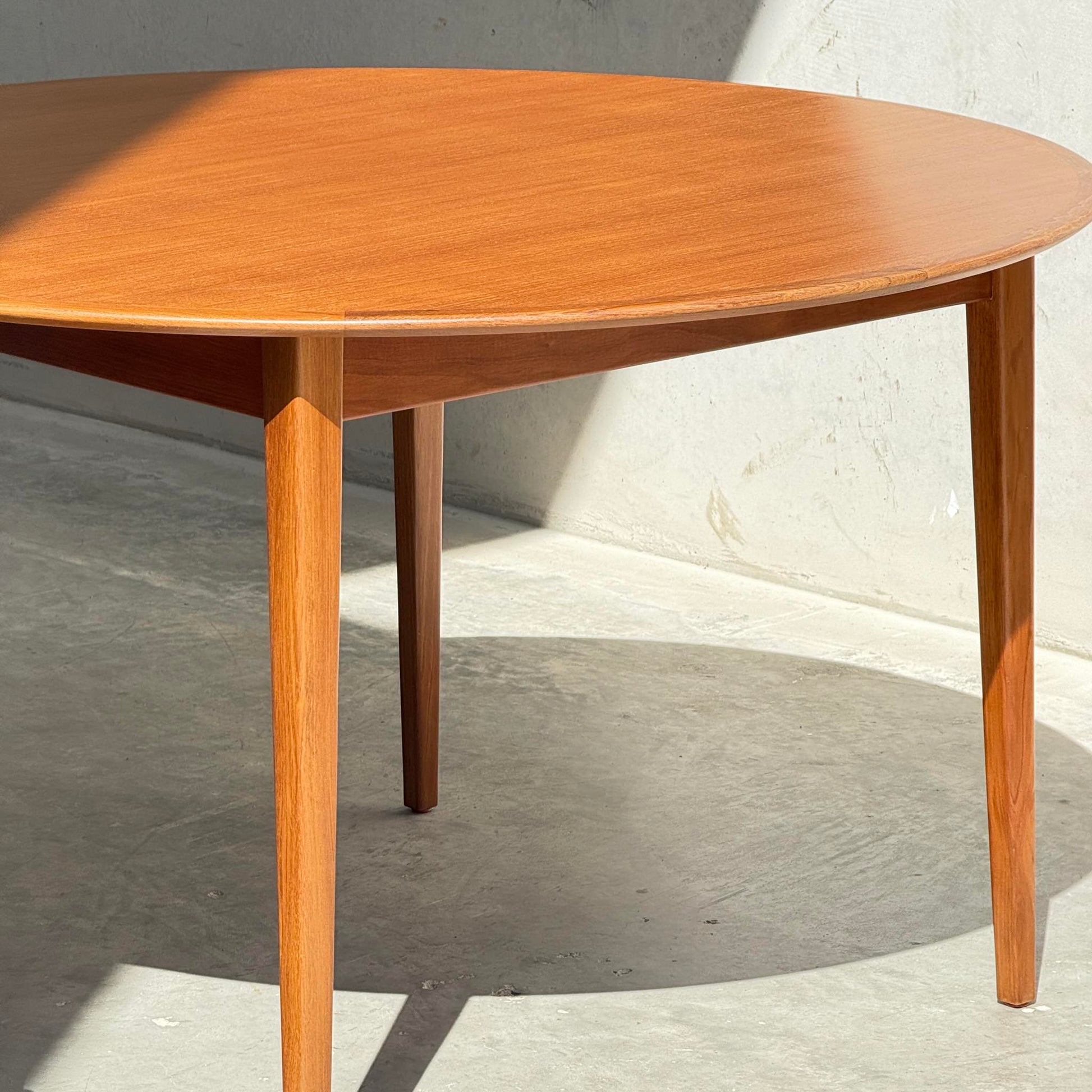 A Fully Restored Noblett Nordic Round MCM Table (Rediscover) with three tapered legs stands on a smooth concrete floor, its lacquered finish catching sunlight, set against a plain light-colored wall.