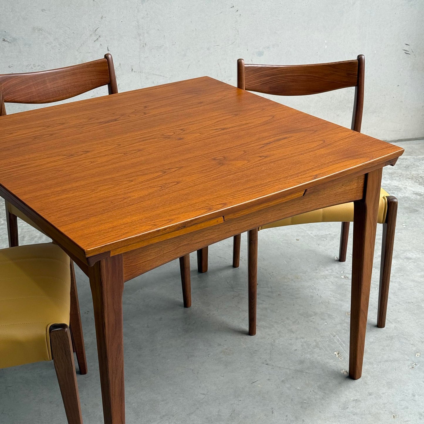 A Fully Restored Fler 64 Square Extension Table (Rediscover) with clean lines is paired with three matching chairs featuring yellow seat cushions, set on a gray concrete floor against a light-colored wall.