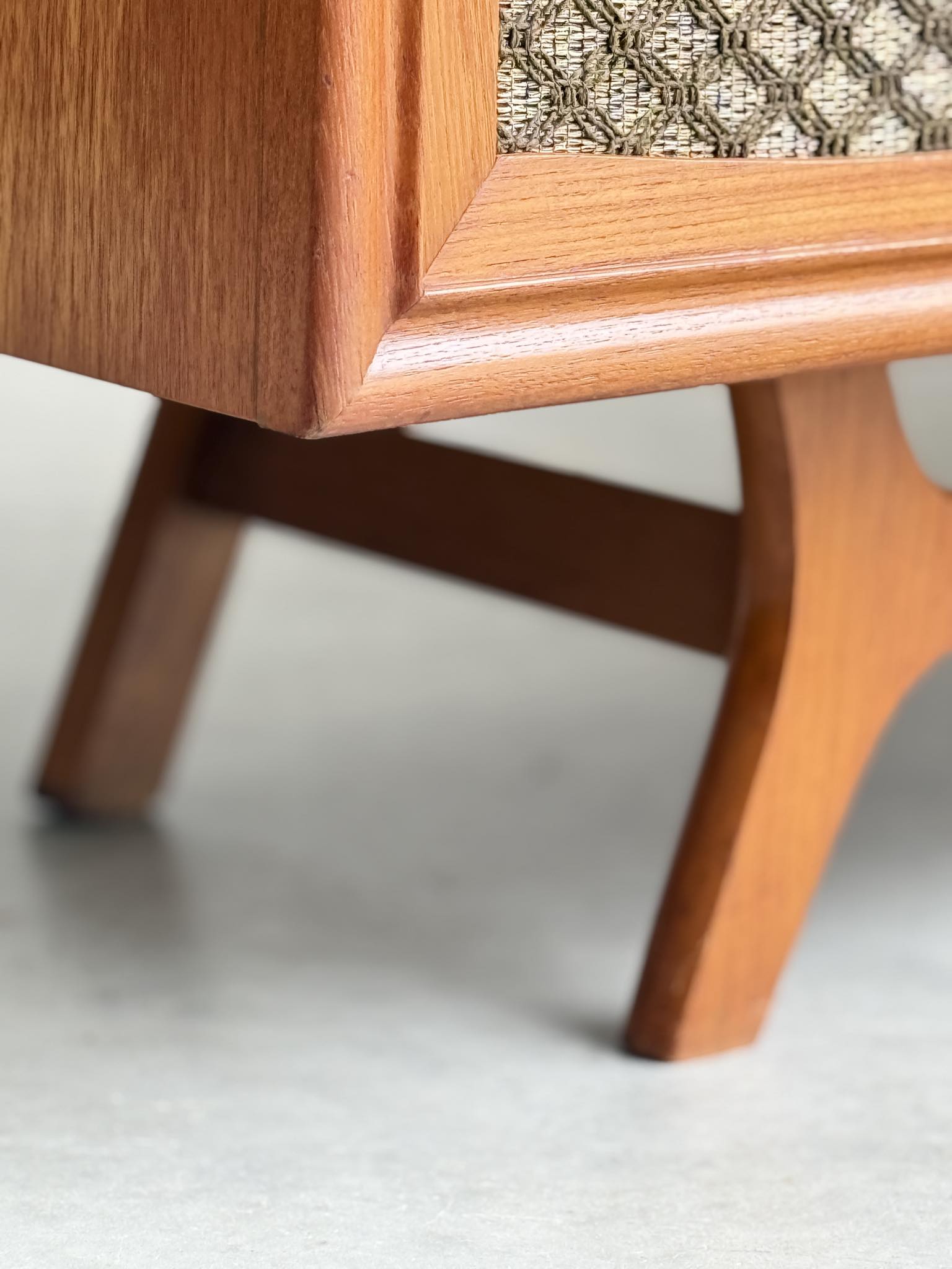Close-up of the General Electric Credenza - Spacemaker 250’s book-matched teak leg, highlighting angled lines and a textured woven fabric panel above a smooth concrete floor.