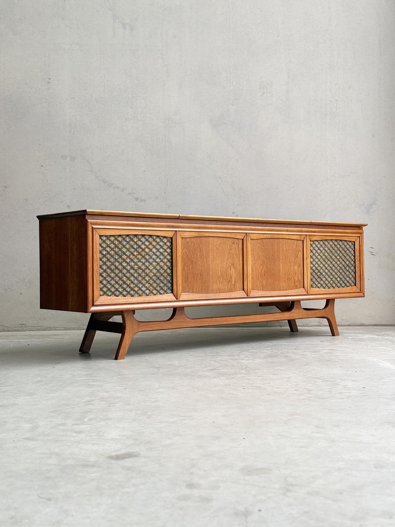 The General Electric Credenza - Spacemaker 250 is a fully converted teak TV/entertainment console with book-matched teak, mid-century modern style, angled legs, and mesh end panels, evoking 1960s design against a plain concrete backdrop.