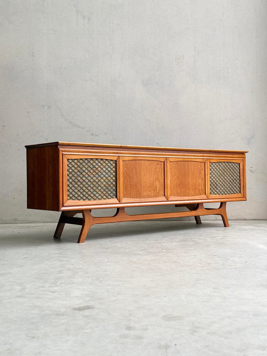 The General Electric Credenza - Spacemaker 250 is a fully converted teak TV/entertainment console with book-matched teak, mid-century modern style, angled legs, and mesh end panels, evoking 1960s design against a plain concrete backdrop.