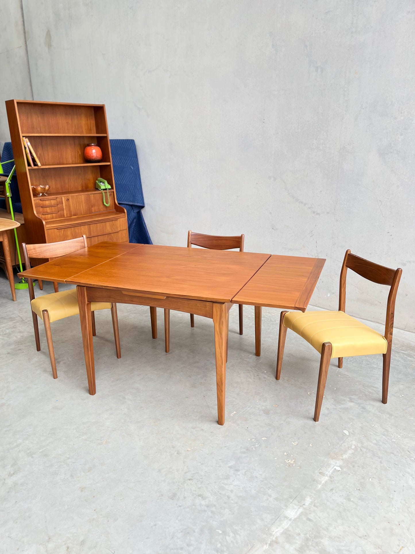A Fully Restored Fler 64 Square extension Table (Rediscover), three wooden chairs with yellow seats, and a matching hutch make up this mid-century modern dining set, displayed elegantly against a plain concrete wall.