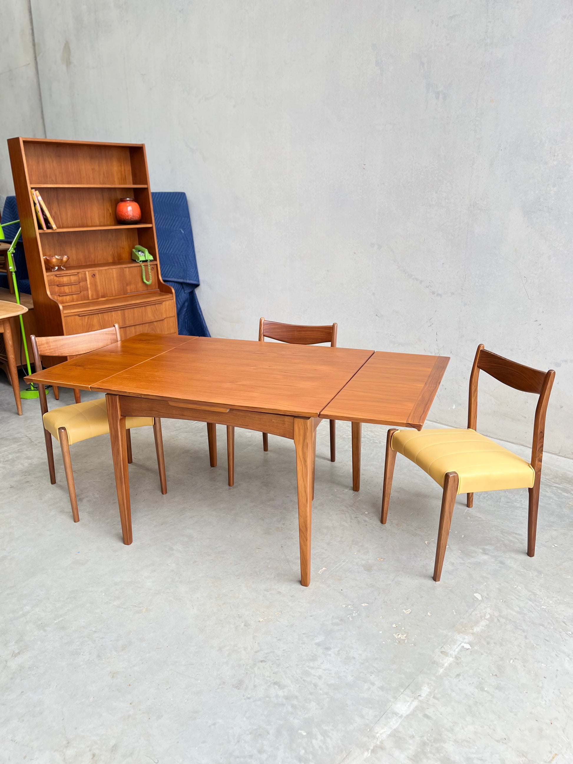 A Fully Restored Fler 64 Square extension Table (Rediscover), three wooden chairs with yellow seats, and a matching hutch make up this mid-century modern dining set, displayed elegantly against a plain concrete wall.
