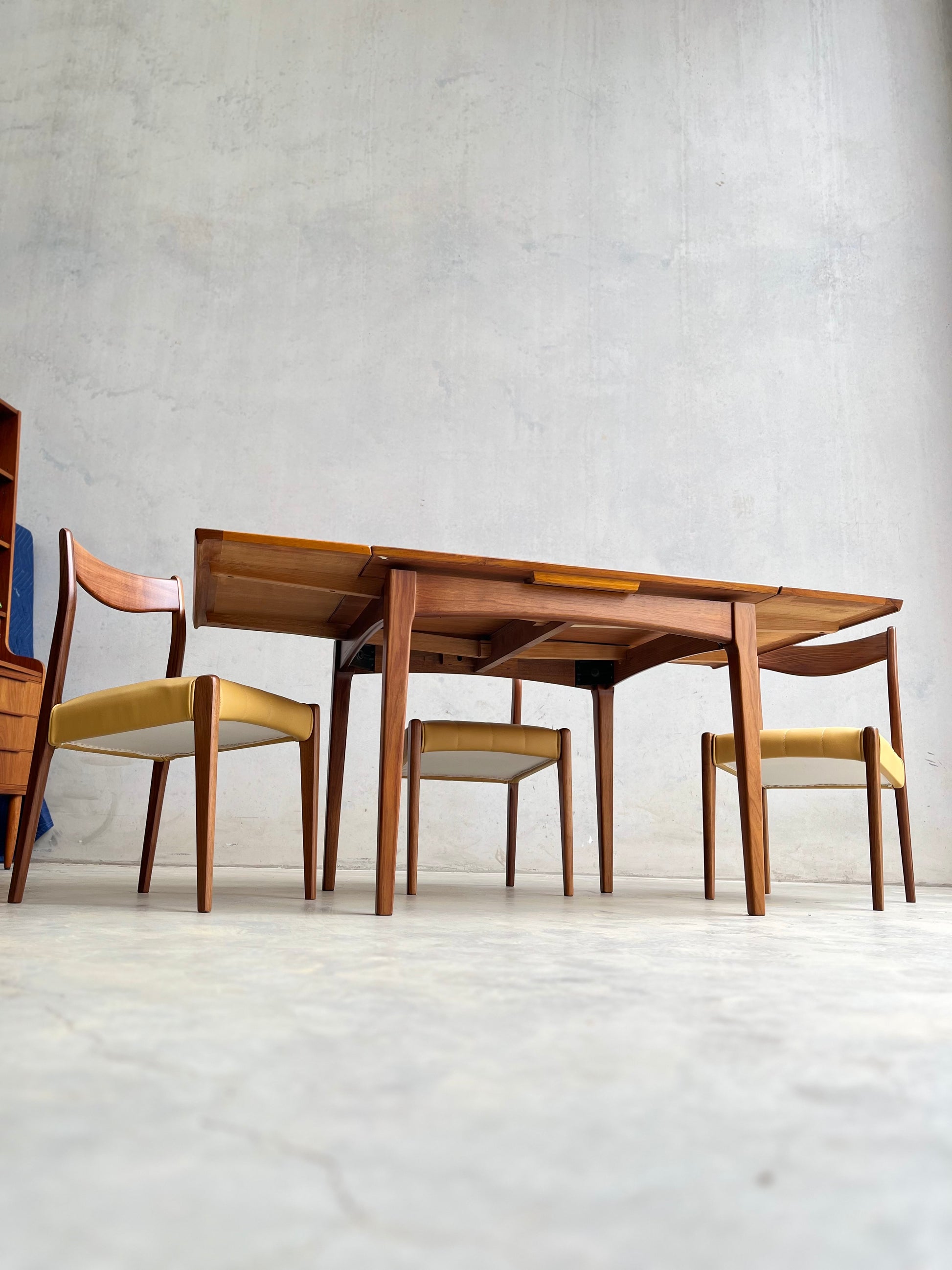 A Fully Restored Fler 64 Square extension Table (Rediscover) by Fred Lowen, paired with four wooden chairs with light yellow cushions, sits against a plain light wall and concrete floor in this mid-century modern dining setup.