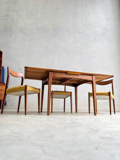 A Fully Restored Fler 64 Square extension Table (Rediscover) by Fred Lowen, paired with four wooden chairs with light yellow cushions, sits against a plain light wall and concrete floor in this mid-century modern dining setup.