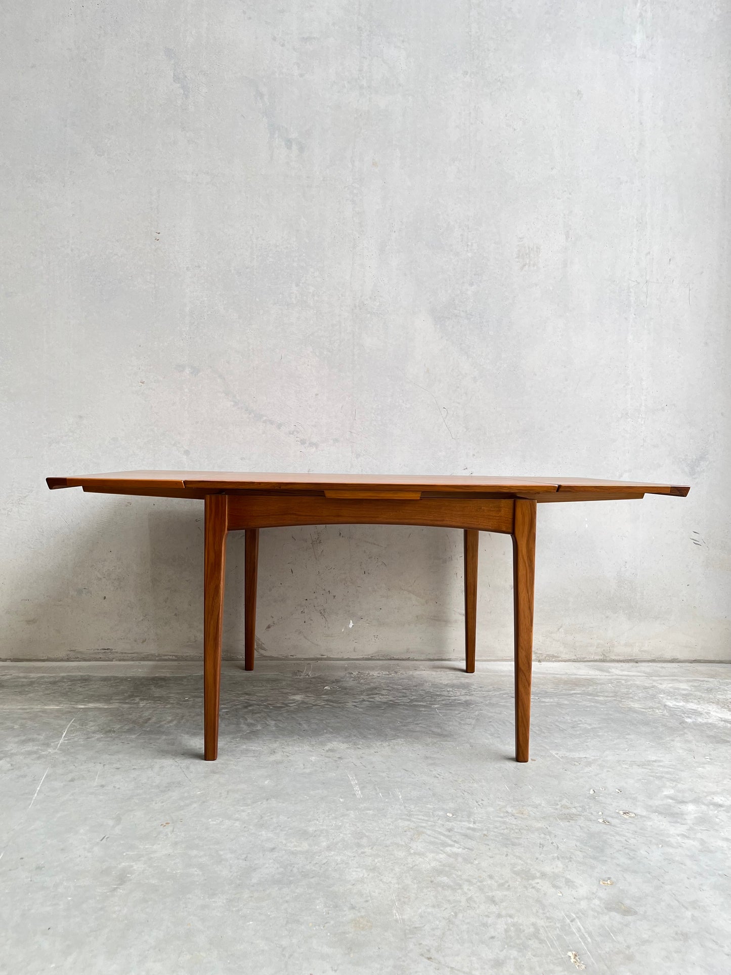 The Fully Restored Fler 64 Square Extension Table (Rediscover) showcases clean lines and tapered legs with extended leaves, inspired by Fred Lowen’s mid-century design, set against a light grey concrete wall and floor.