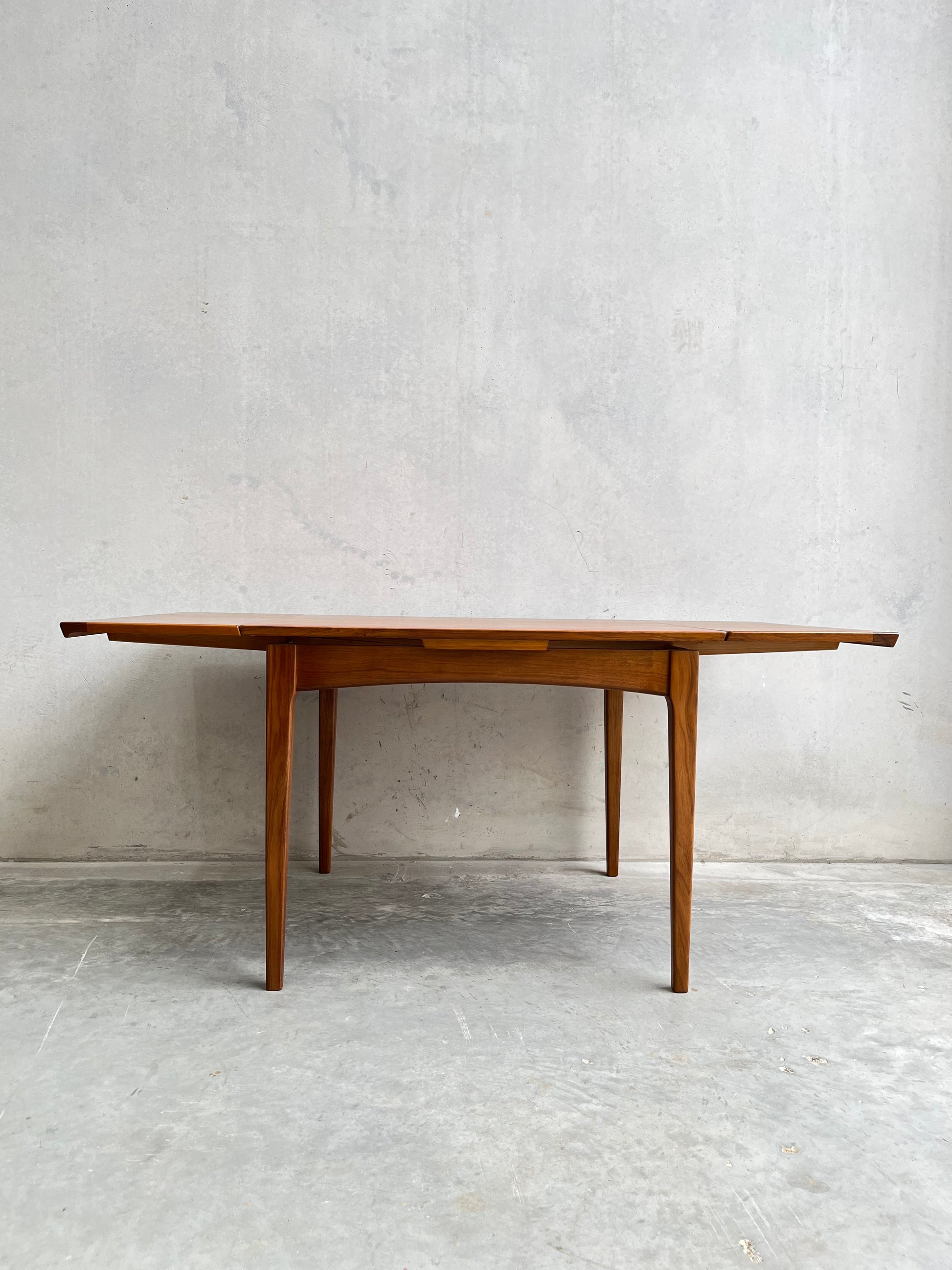 The Fully Restored Fler 64 Square Extension Table (Rediscover) showcases clean lines and tapered legs with extended leaves, inspired by Fred Lowen’s mid-century design, set against a light grey concrete wall and floor.