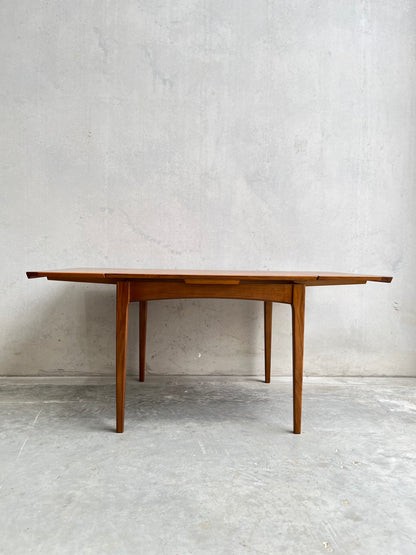 The Fully Restored Fler 64 Square Extension Table (Rediscover) showcases clean lines and tapered legs with extended leaves, inspired by Fred Lowen’s mid-century design, set against a light grey concrete wall and floor.