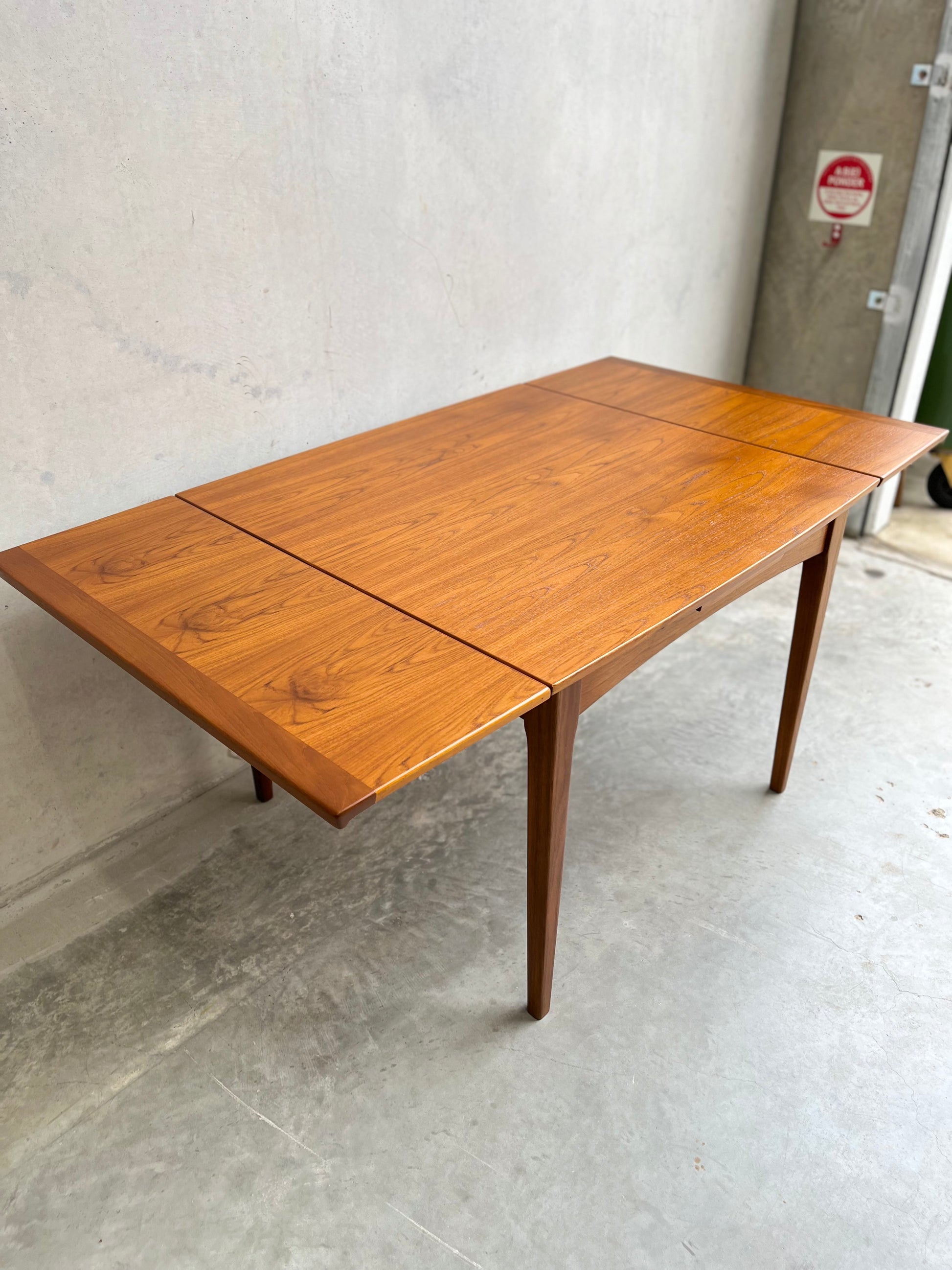 A Fully Restored Fler 64 Square Extension Table (Rediscover) sits against a light concrete wall and floor, with extendable leaves at both ends, near a metal door with a red sign—a perfect mid-century modern dining table setting.