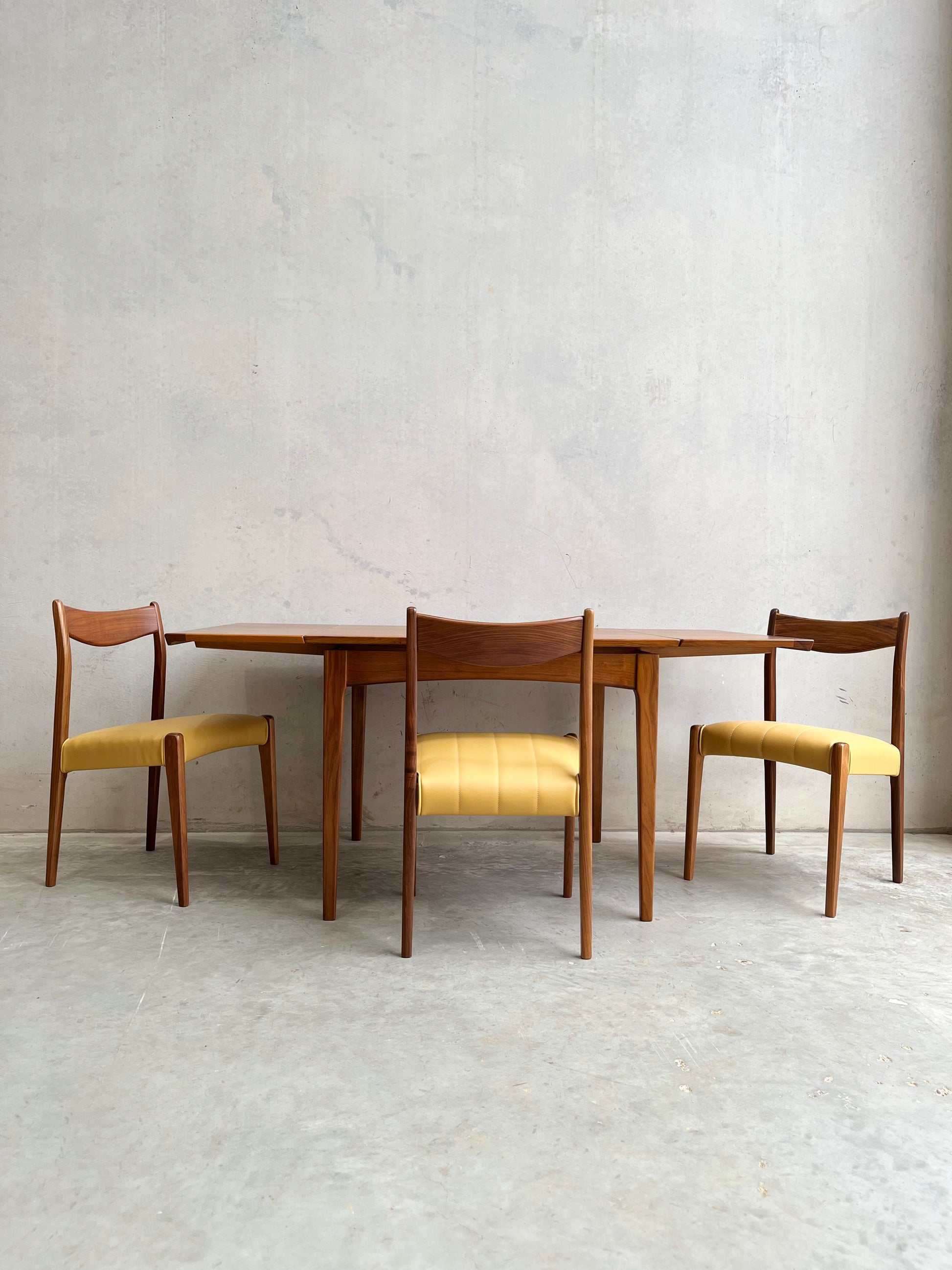 A fully restored Fler 64 Square extension table (Rediscover) with three wooden chairs featuring yellow upholstered seats, set against a plain light gray concrete wall and floor.