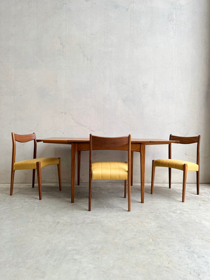 A fully restored Fler 64 Square extension table (Rediscover) with three wooden chairs featuring yellow upholstered seats, set against a plain light gray concrete wall and floor.