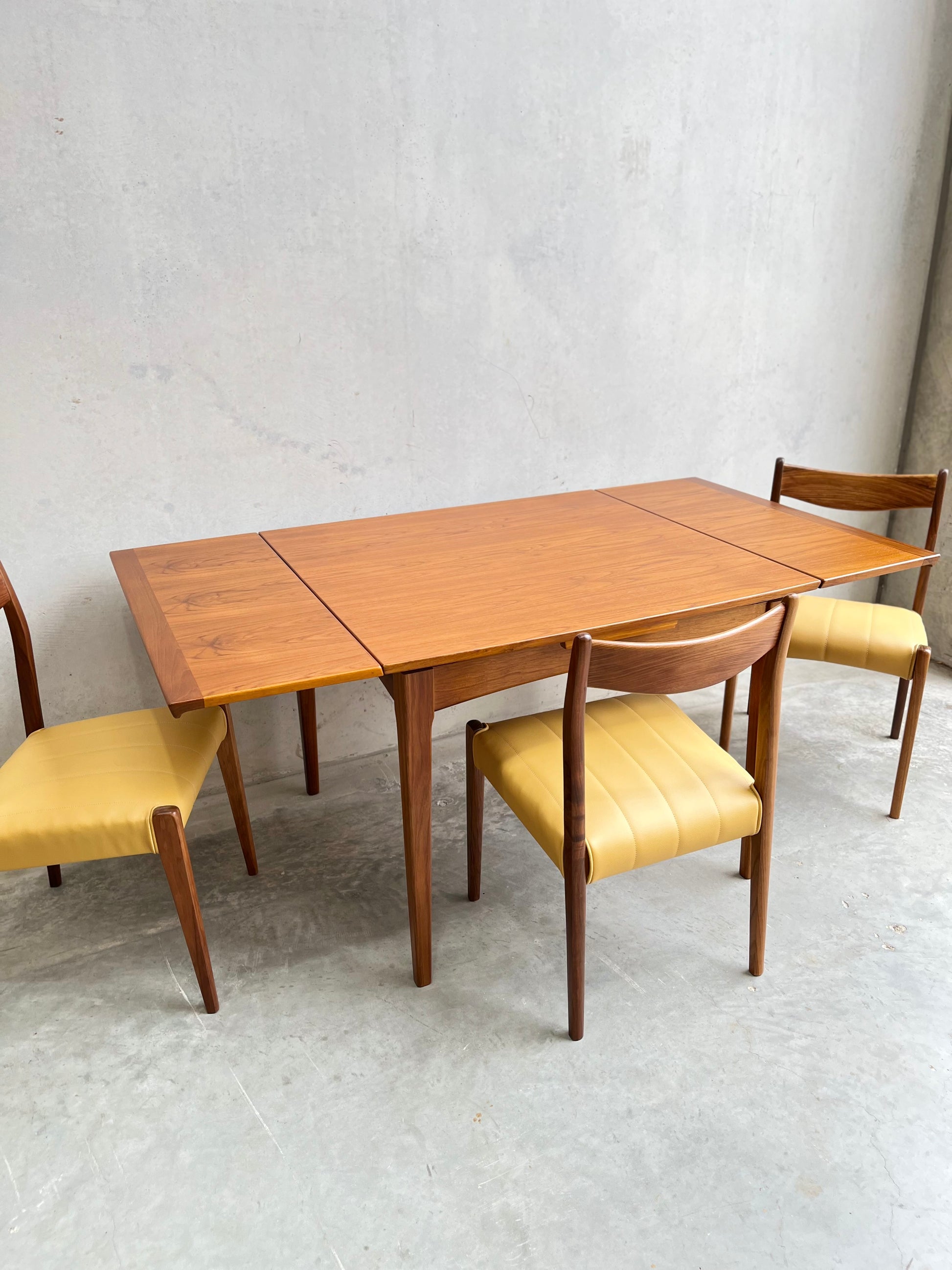 A fully restored Fler 64 Square extension table (Rediscover) with clean lines and three matching wooden chairs featuring yellow upholstered seats, set on a gray concrete floor against a plain light gray wall.