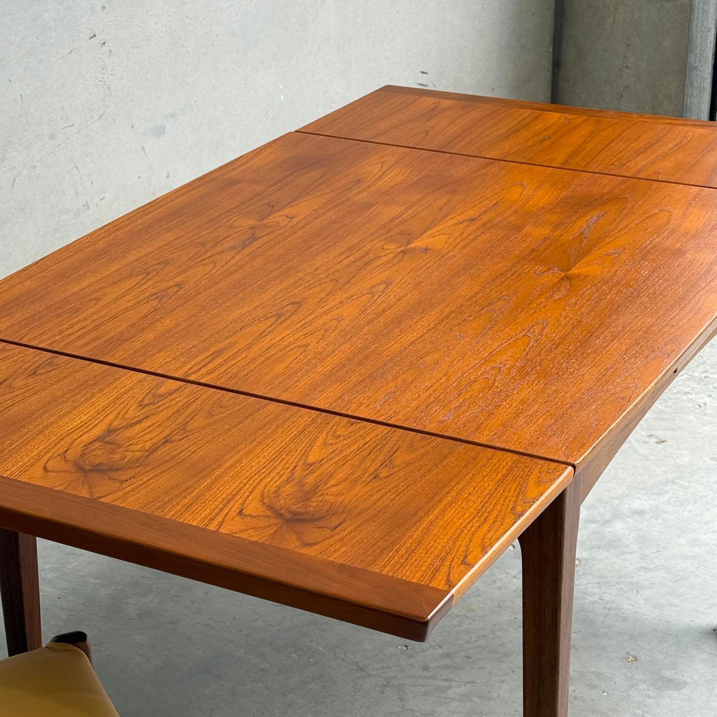 A Fully Restored Fler 64 Square Extension Table (Rediscover) with a brown wood finish and grain detail sits near a gray concrete wall. A yellow chair appears in the lower left, evoking classic mid-century modern dining style.