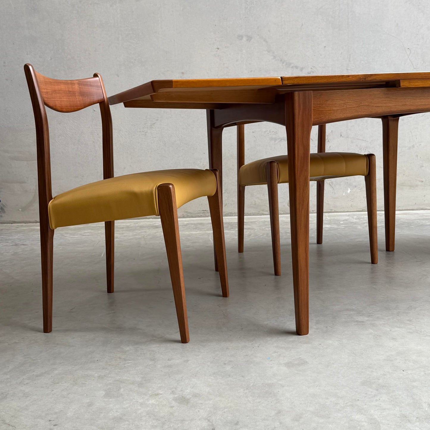 Two mid-century modern wooden chairs with light brown cushions are placed at a fully restored Fler 64 Square extension table (Rediscover), set against a plain gray concrete wall and floor.